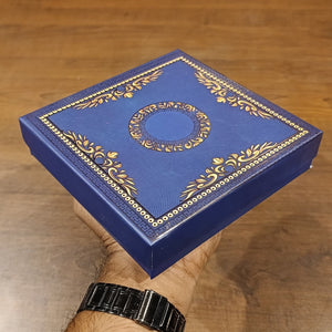 A hand holding a square blue presentation box with ornate gold foil detailing and a circular emblem on the lid, set against a wooden background.