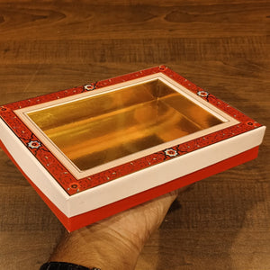 A hand holding a rectangular gift box with a transparent window showing a gold interior, featuring a red patterned border and white sides, set against a wooden background.