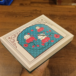 A decorative rectangular gift box with a cream-colored border and a central turquoise panel featuring pink lotus flowers, white birds, and wave patterns, resting on a wooden table with a hand holding it.