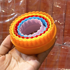 A hand holding a set of nested round plastic ring molds in yellow, orange, red, blue, and pink, photographed against a reflective brown background.