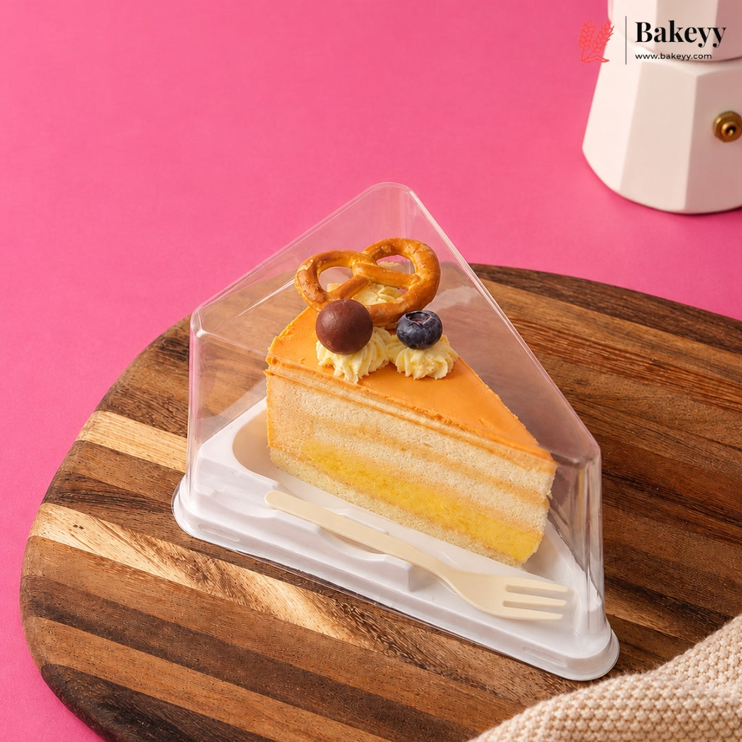 A triangular clear plastic cake display box containing a slice of layered cake with whipped cream, a pretzel, and blueberries, placed on a wooden board against a pink background. The Bakeyy logo appears in the top right corner.