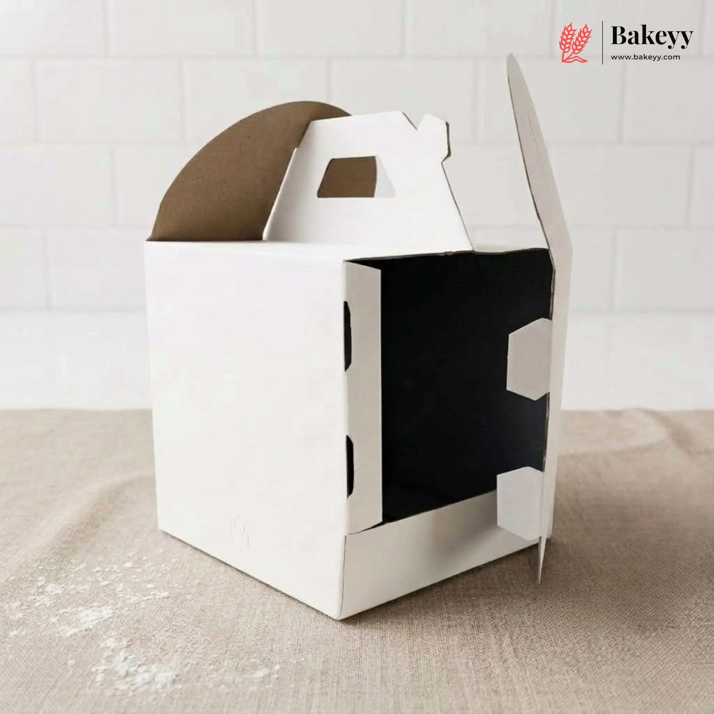 White cardboard cake box with a brown cake inside on a textured surface, branded 'Bakeyy'.