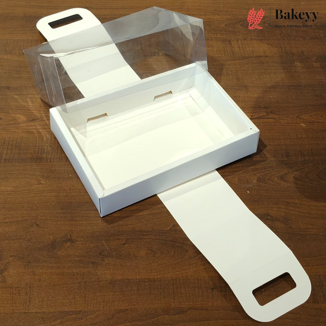 A white rectangular bakery box with a clear plastic lid and white paper flaps, shown on a brown wooden tabletop. The Bakeyy logo is visible in the top right corner.