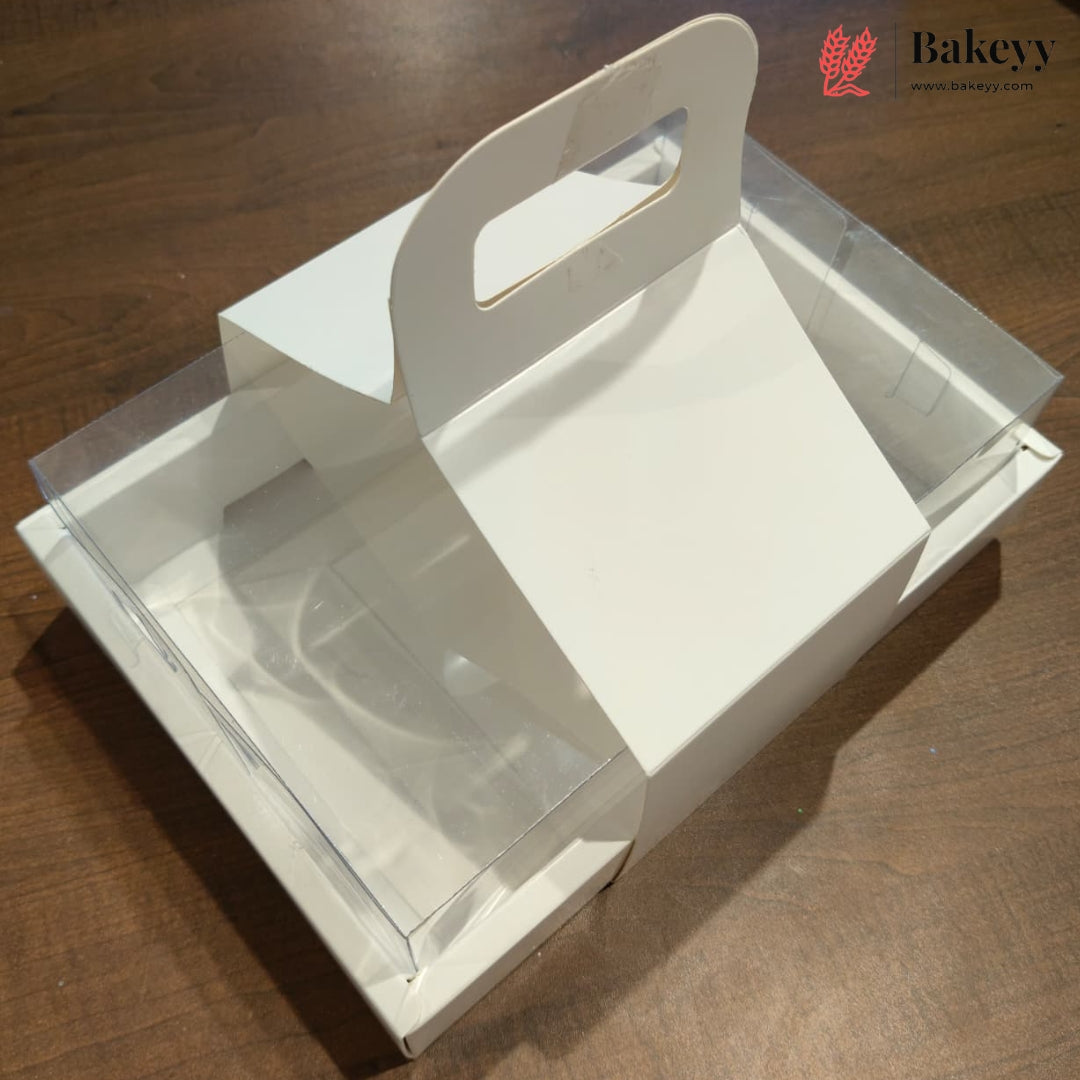 A white cardboard bakery or gift box with a built-in handle cutout and clear plastic side windows, shown on a wooden tabletop. A Bakeyy logo watermark is visible in the top right corner.