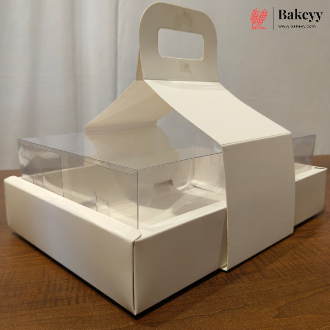 A white cardboard bakery display box with a built-in handle and clear plastic window panels, shown on a wooden tabletop against a light curtain background. A Bakeyy logo watermark is visible in the top right corner.