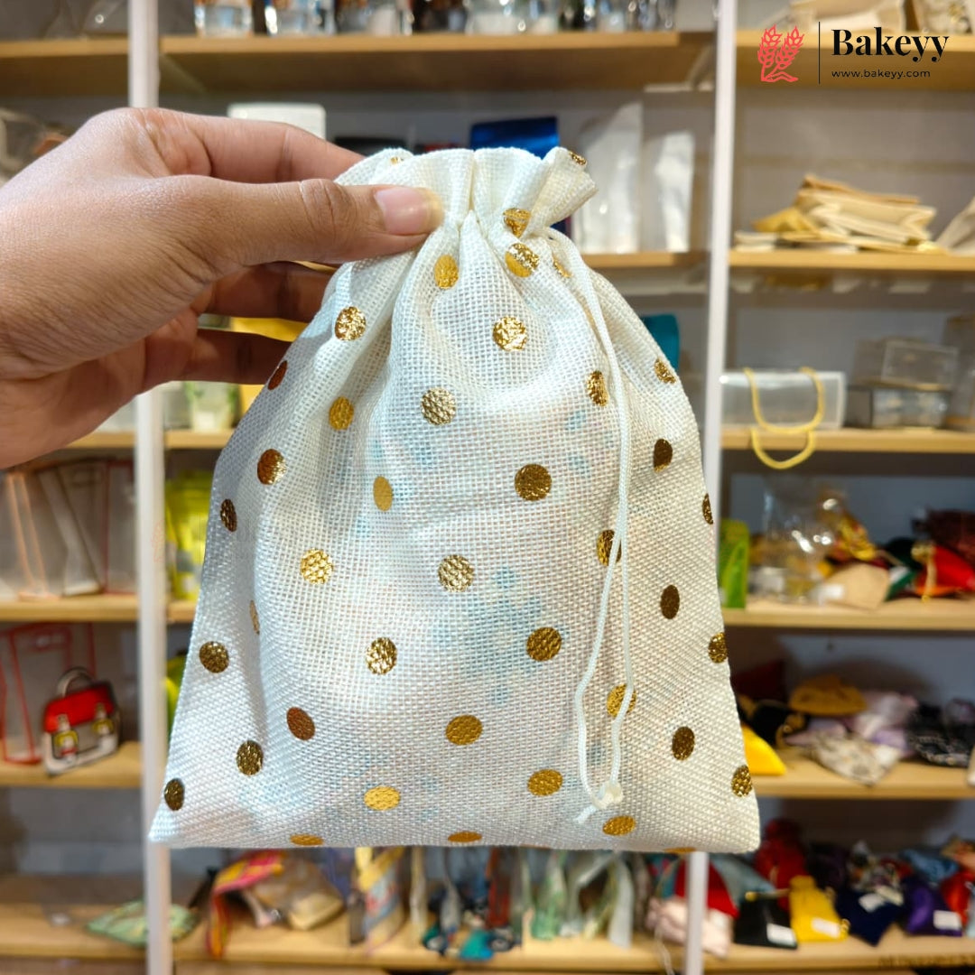 A hand holds a small drawstring pouch made of white organza fabric with scattered gold polka dots. The background shows a retail store shelf filled with various items.