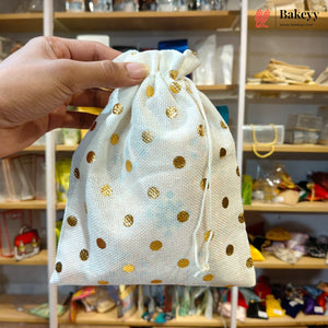 A hand holds a small drawstring pouch made of white organza fabric with scattered gold polka dots. The background shows a retail store shelf filled with various items.