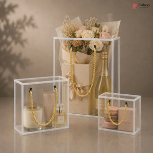 Clear display boxes with gold handles containing a bottle of wine, flowers, and other items on a neutral background.