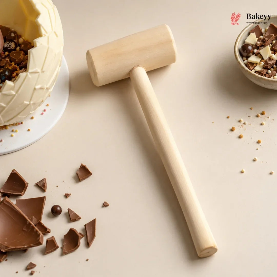 A wooden cake smash hammer with a smooth cylindrical head and long handle, styled on a beige surface with chocolate shards, a partially broken cream cake, and a small bowl of chocolate pieces. Bakeyy logo appears in the top right corner.