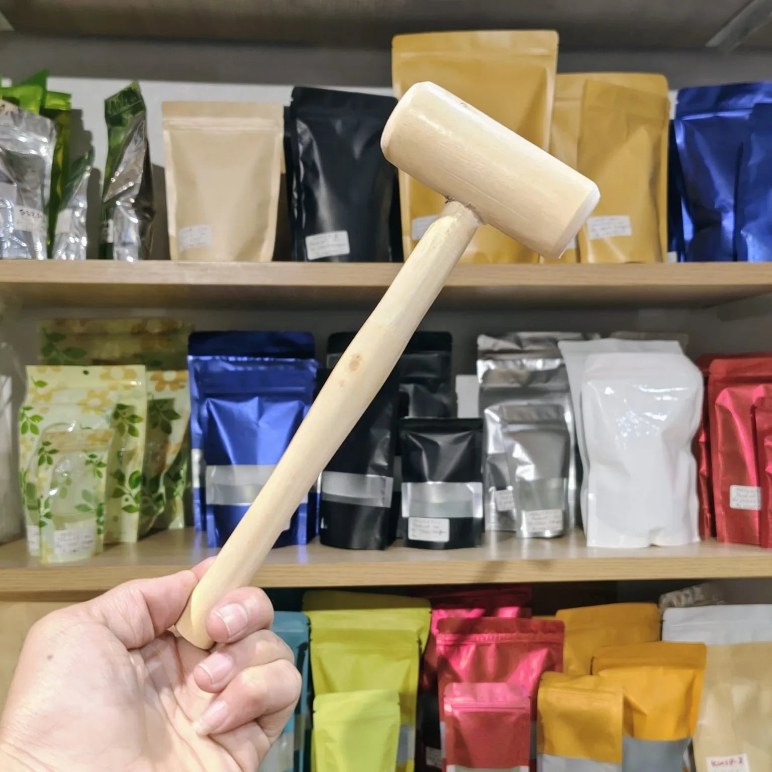 A hand holding a light-colored wooden mallet against a background of shelves stocked with various colorful pouches of tea and coffee.