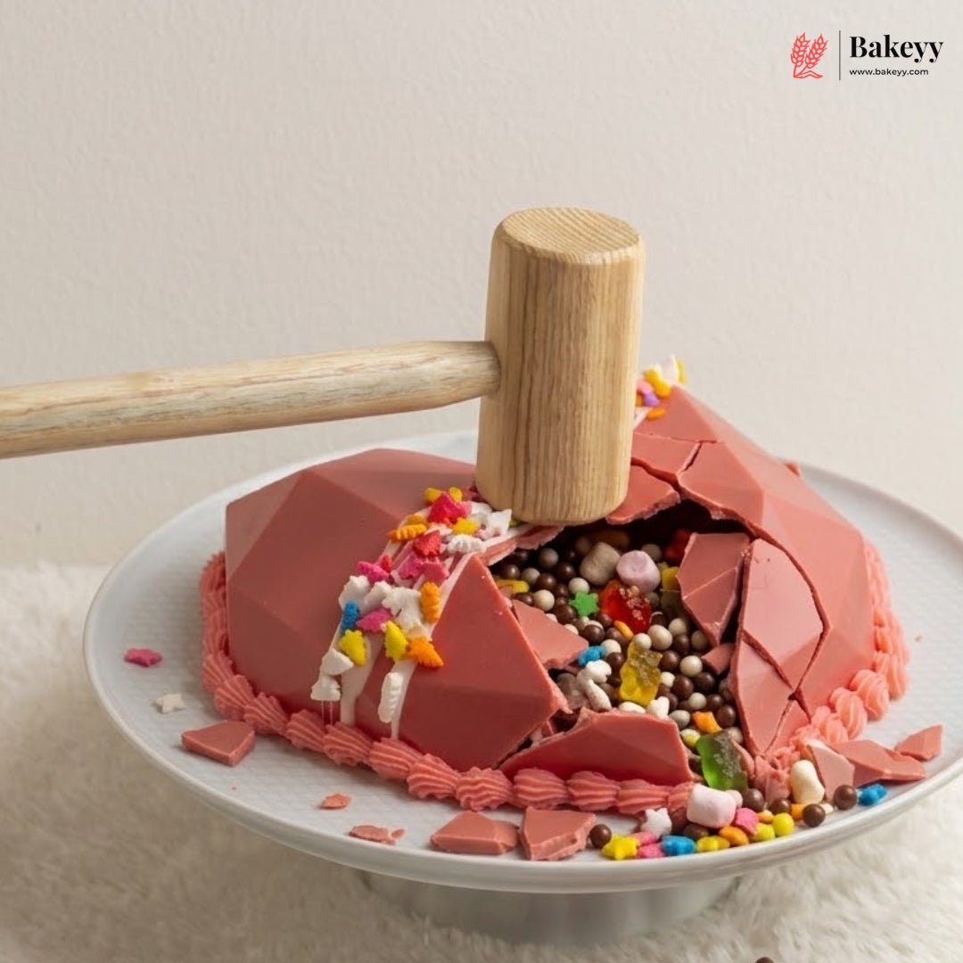 A pink chocolate cake on a white plate with a wooden mallet resting on top, smashing the cake to reveal colorful candy and chocolate pieces inside. The Bakeyy logo is visible in the top right corner.