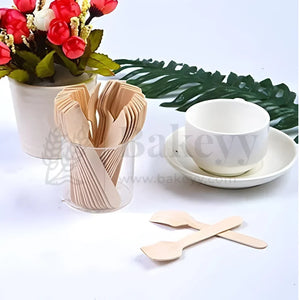 Wooden cutlery spoons in a clear cup on white table with cup, saucer, flowers, at Bakeyy.com