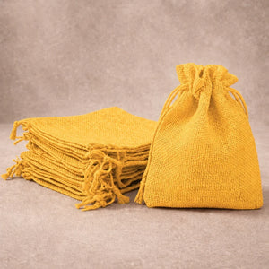 A stack of mustard yellow burlap drawstring gift bags with fringe, with one bag standing open on the right, set against a neutral textured gray background.