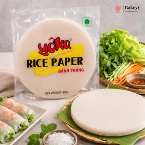 A package of Yoka Rice Paper (Bánh Tráng) displayed alongside fresh spring rolls, lettuce, herbs, and sliced carrots on a light gray surface.