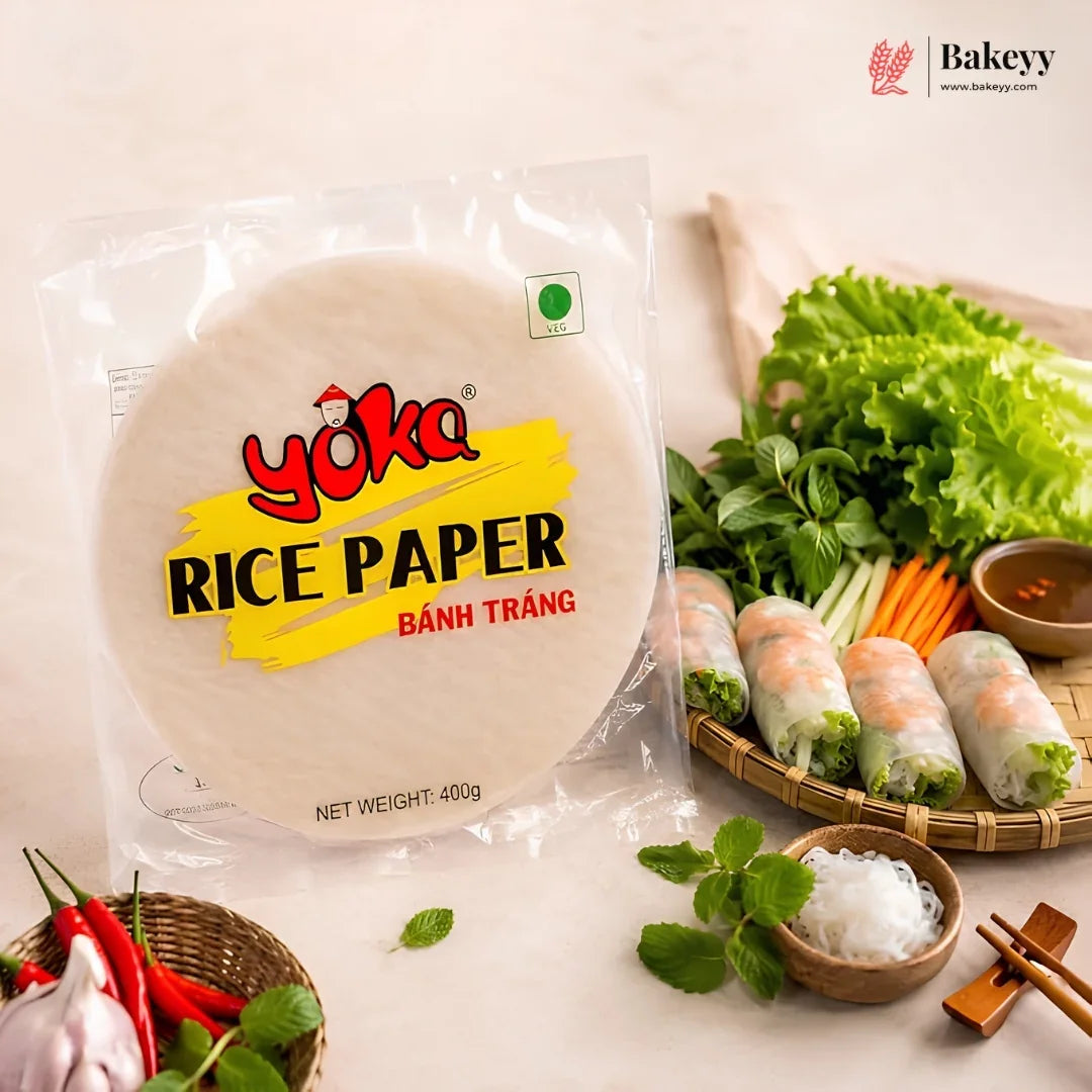 A package of Yoka Rice Paper (Bánh Tráng) displayed next to fresh spring roll ingredients, including lettuce, herbs, carrots, garlic, and prepared spring rolls on a light beige background.