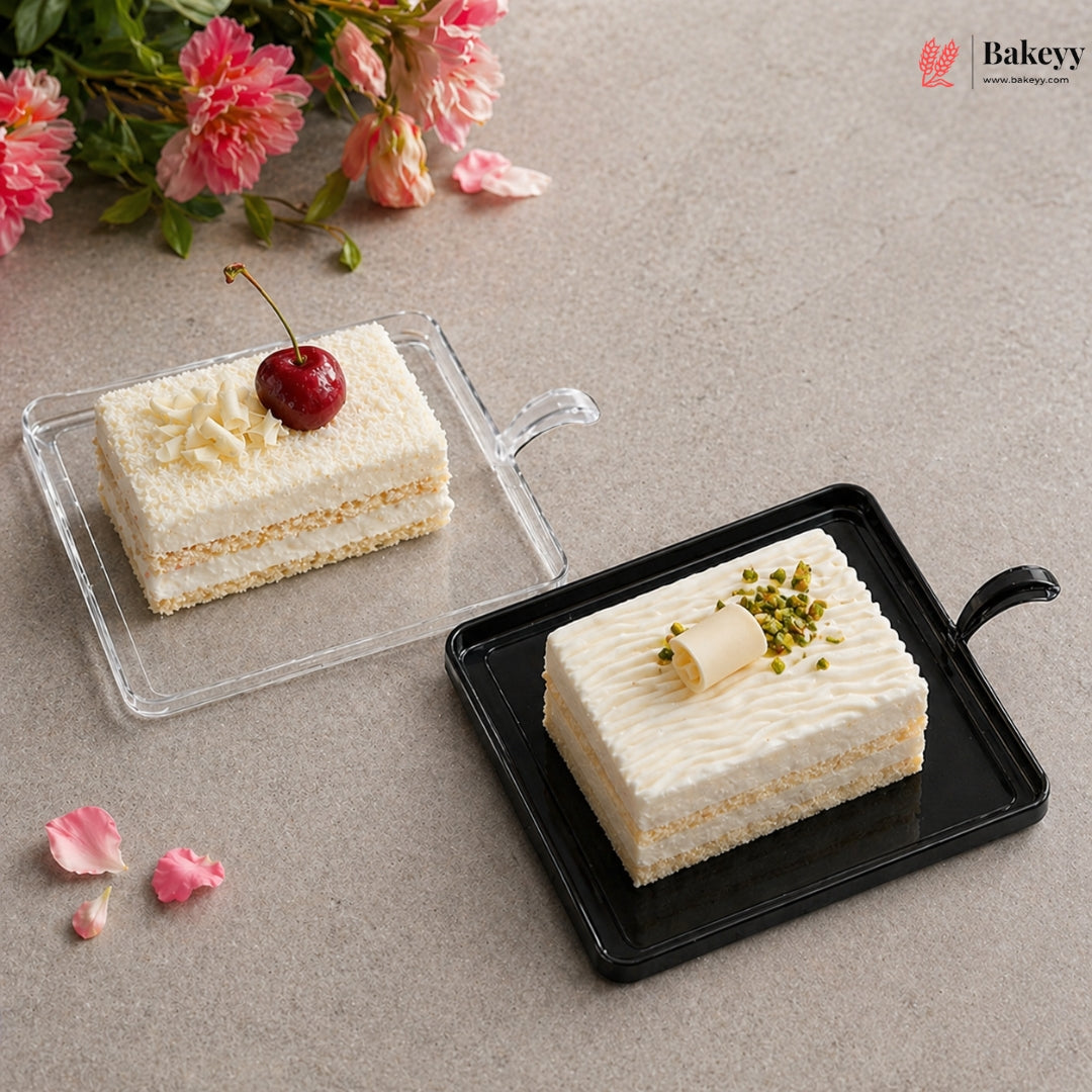 Two slices of cake on transparent and black trays with pink flowers in the background, featuring the Bakeyy brand.