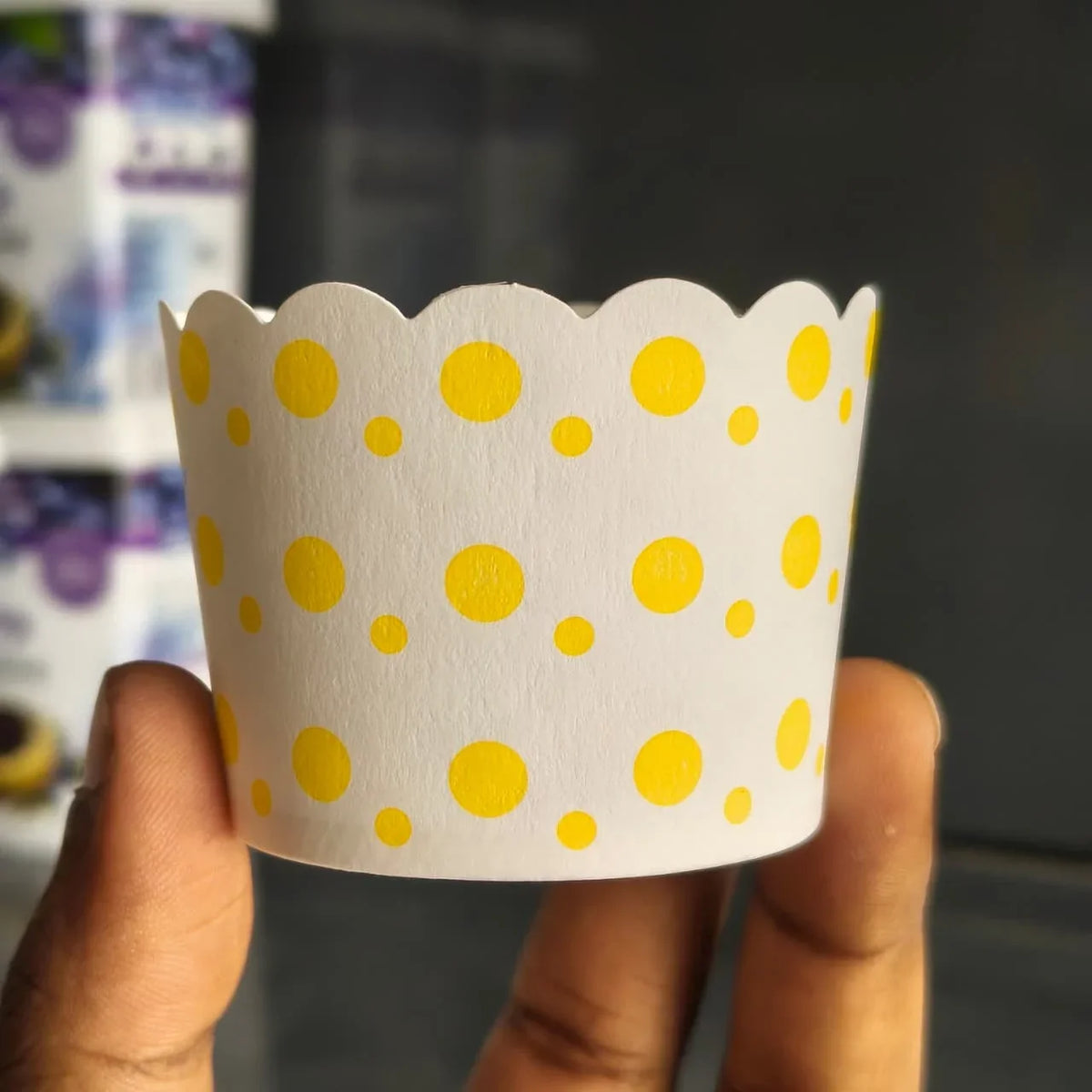 A hand holding a white paper cupcake liner decorated with yellow polka dots, with a blurred background.