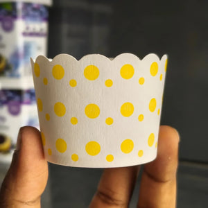 A hand holding a white paper cupcake liner decorated with yellow polka dots, with a blurred background.