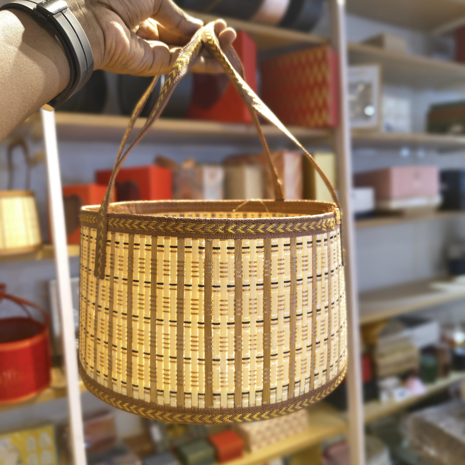 A hand holds a small woven basket with two handles in a shop setting. The basket has a natural light wood body with dark brown woven bands and decorative geometric patterns near the rim and base.