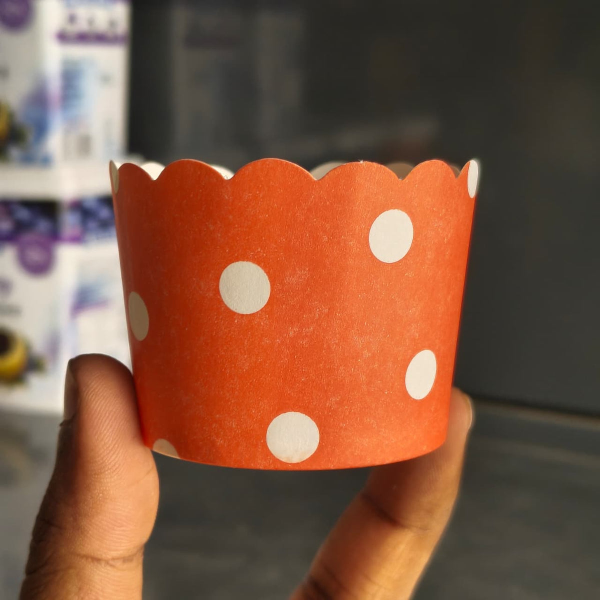 An orange paper cupcake liner with white polka dots and a scalloped edge, held by a hand against a blurred background.