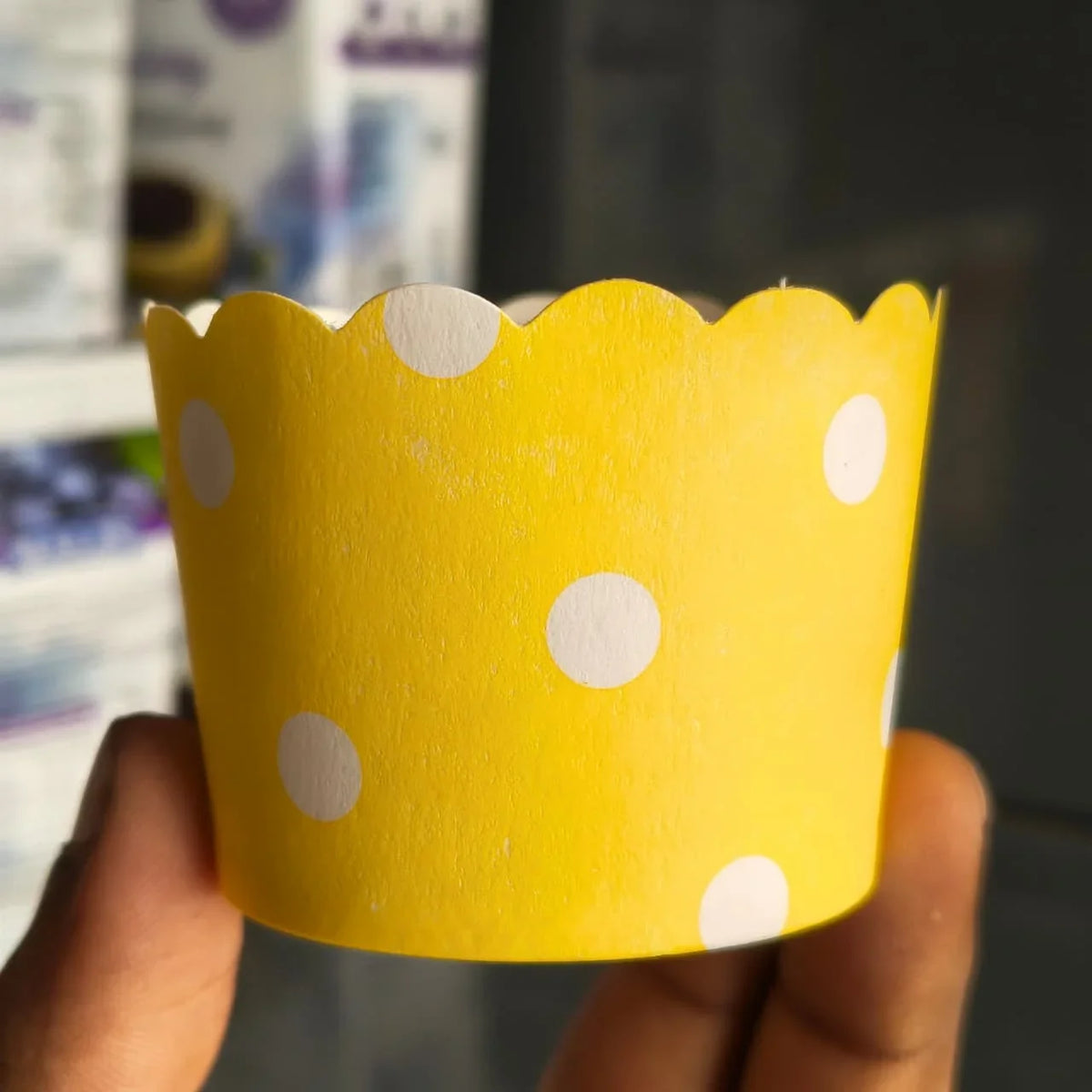 A hand holding a yellow paper cupcake liner with white polka dots, set against a blurred background.