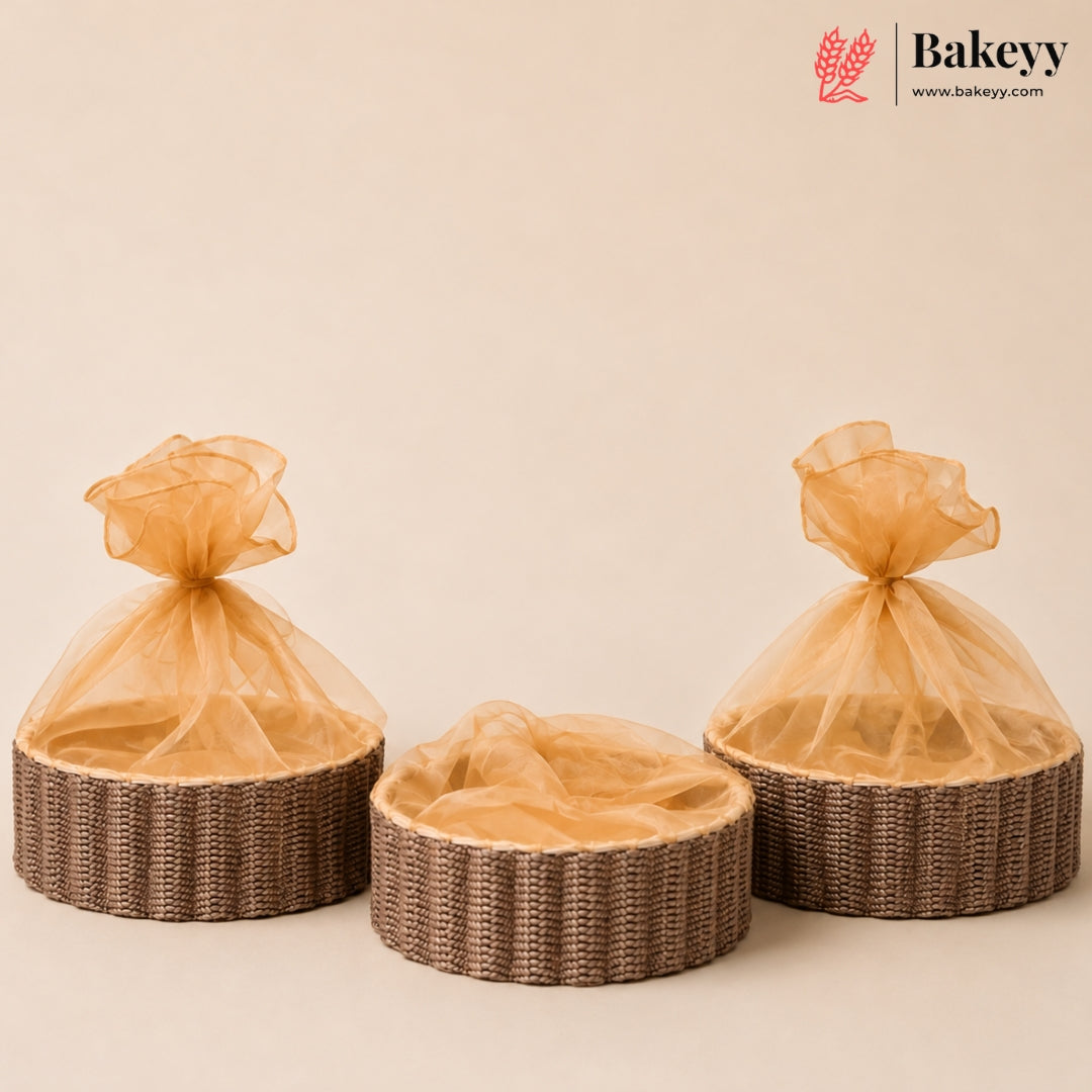 Three round woven wicker baskets with sheer champagne-colored organza bags tied at the top, arranged on a warm beige background. A Bakeyy logo is visible in the top right corner.