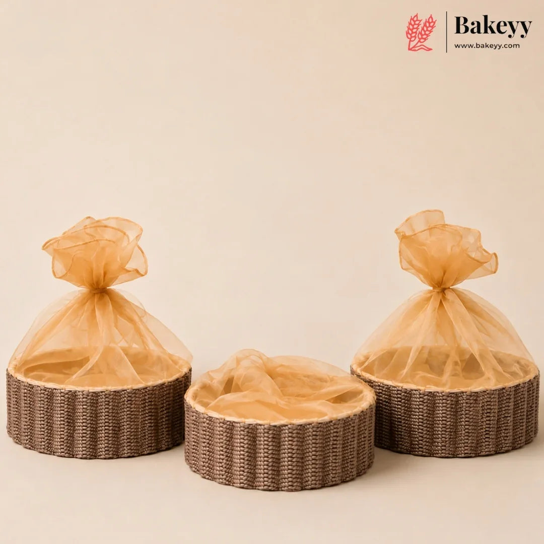 Three round woven wicker baskets with sheer champagne-colored organza bags tied at the top, arranged on a warm beige background. A Bakeyy logo is visible in the top right corner.