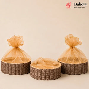 Three round woven wicker baskets with sheer champagne-colored organza bags tied at the top, arranged on a warm beige background. A Bakeyy logo is visible in the top right corner.