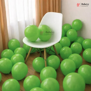 Bright green latex balloons scattered across a wooden floor and draped around a white molded chair in an indoor room with curtains.