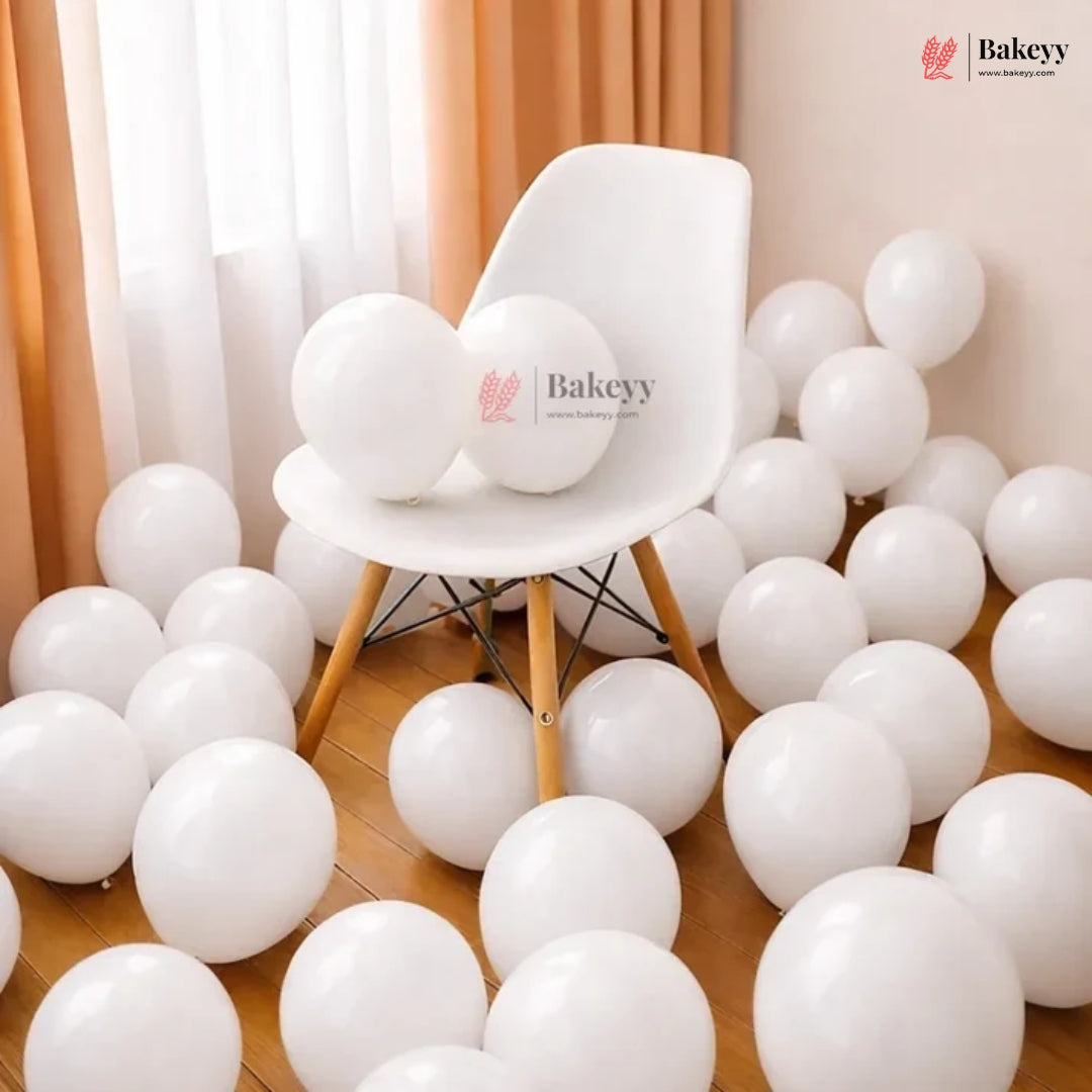 White round party balloons scattered across a wooden floor, with two balloons resting on a white chair in the center. The scene is styled with peach curtains and a warm beige wall.