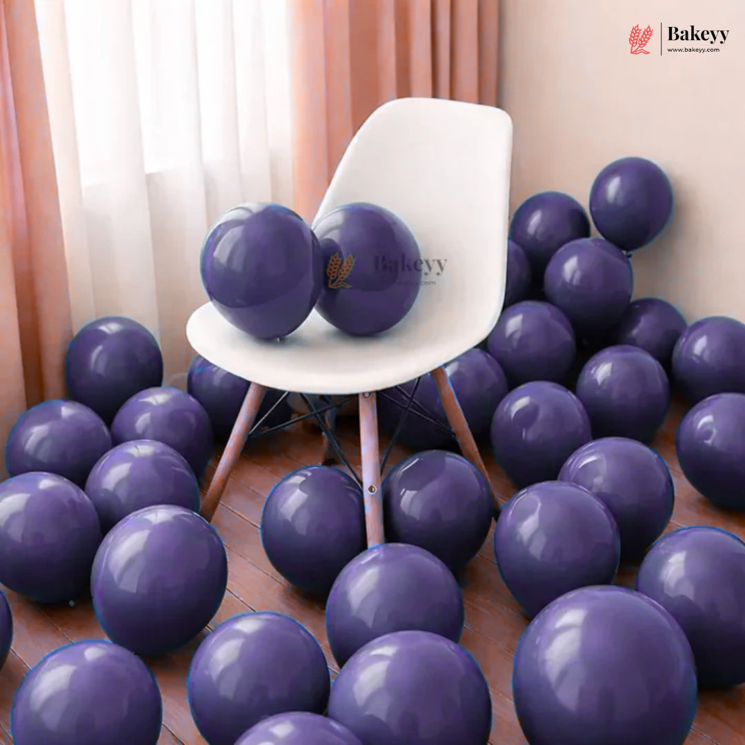 A room scene filled with glossy dark purple balloons, with two balloons resting on a white chair. The background includes peach curtains and a light-colored wall, and a Bakeyy logo watermark is visible in the top right.