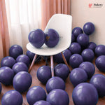 A room scene filled with glossy dark purple balloons, with two balloons resting on a white chair. The background includes peach curtains and a light-colored wall, and a Bakeyy logo watermark is visible in the top right.