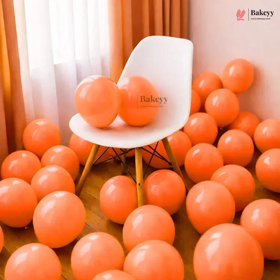 Orange balloons scattered across a wooden floor around a white chair, with two orange balloons resting on the chair. The room has peach walls, sheer white curtains, and orange drapes.