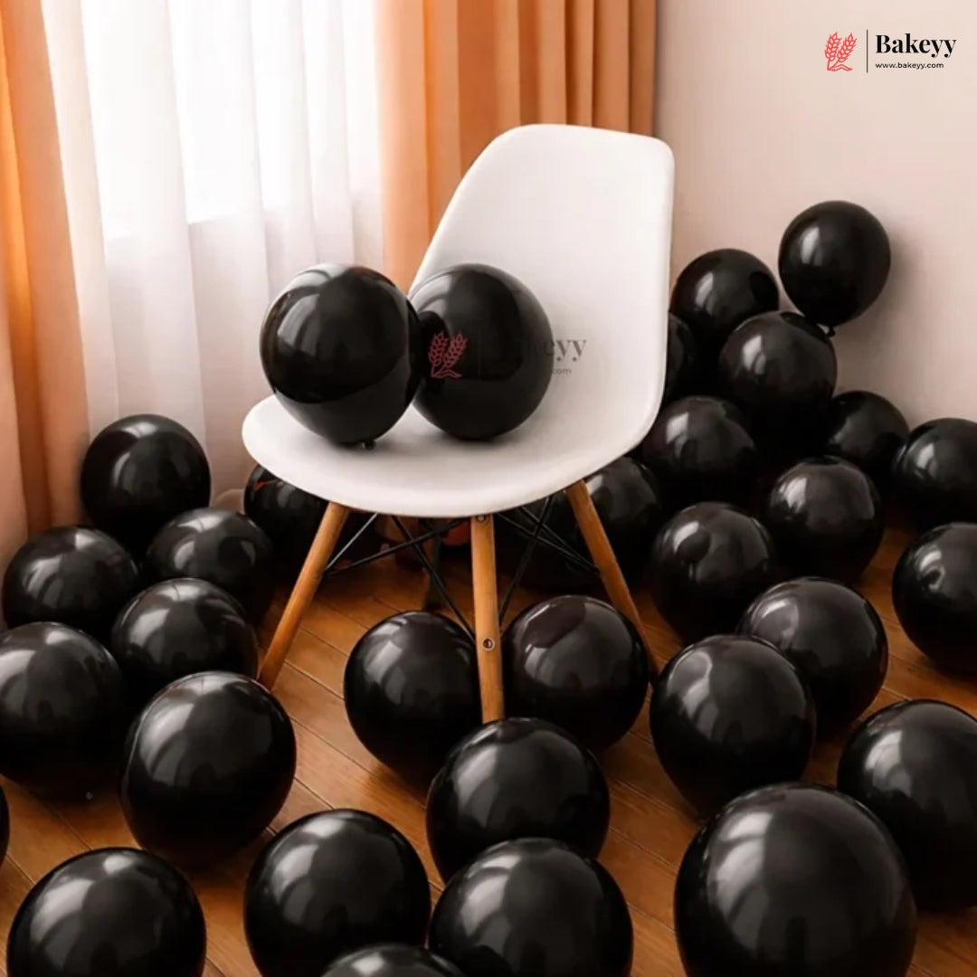 Black round balloons scattered across a wooden floor and draped around a white molded chair in a room with sheer curtains. Two black balloons rest on the chair, and a Bakeeyy logo appears in the top right corner.