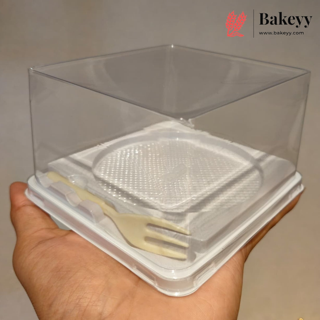 A hand holding a clear plastic bakery display box with a white base and a small beige plastic fork inside, against a plain light background. The Bakeyy logo is visible in the top right corner.