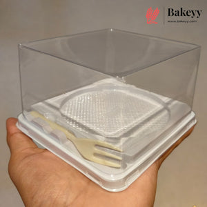 A hand holding a clear plastic bakery display box with a white base and a small beige plastic fork inside, against a plain light background. The Bakeyy logo is visible in the top right corner.