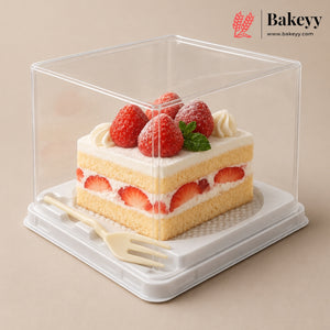 A clear square plastic cake display box with a white base, shown containing a slice of strawberry cake with cream and fresh strawberries on top. A small cream fork rests inside the box.