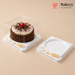 A clear square plastic cake box with a white base holding a chocolate cake topped with cream and a red cherry, shown next to the matching empty lid on a beige background. The Bakeyy logo appears in the top right corner.
