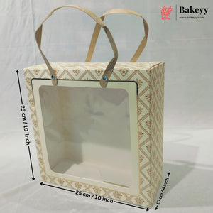 A square bakery gift box with a clear front window and beige paper handles, shown with dimension arrows indicating 20 cm by 20 cm by 10 cm measurements on a white backdrop. A Bakeyy logo appears in the top right.