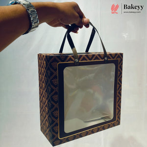 A hand wearing a silver watch holds a rectangular bakery gift box with a clear front window and dark brown handles. The box has a repeating geometric pattern in dark brown and gold.