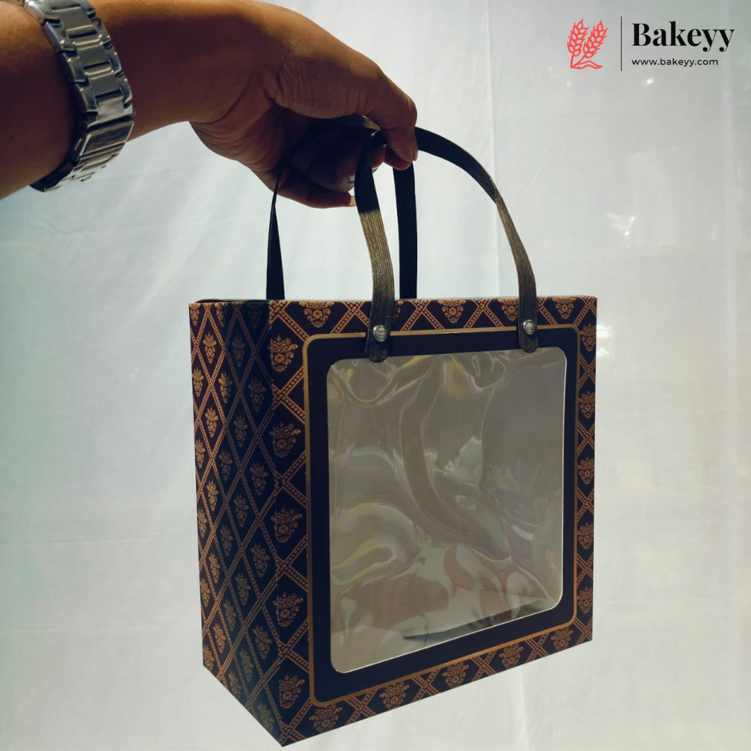 A hand holds a square bakery gift bag with a clear plastic window and dark handles. The bag has a dark navy blue and brown geometric patterned design.