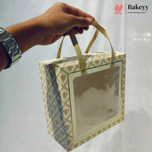 A hand holds a square bakery gift box with a clear front window and gold ribbon handles. The box has a beige and brown geometric pattern.