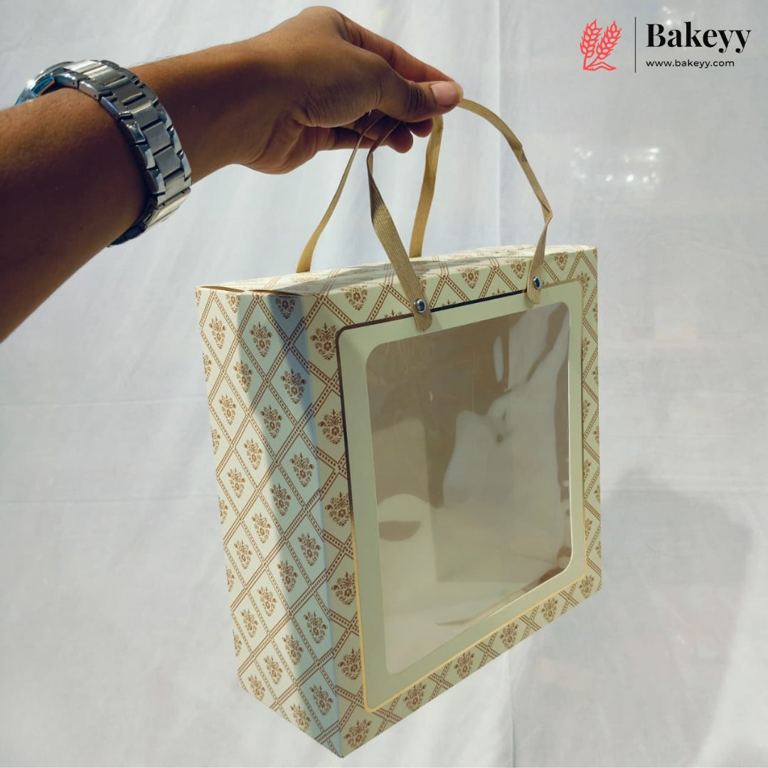 A hand wearing a silver watch holds a square bakery gift bag with a clear window and beige handles. The bag has a cream and gold geometric pattern, set against a white draped backdrop with a Bakeyy logo in the top right corner.