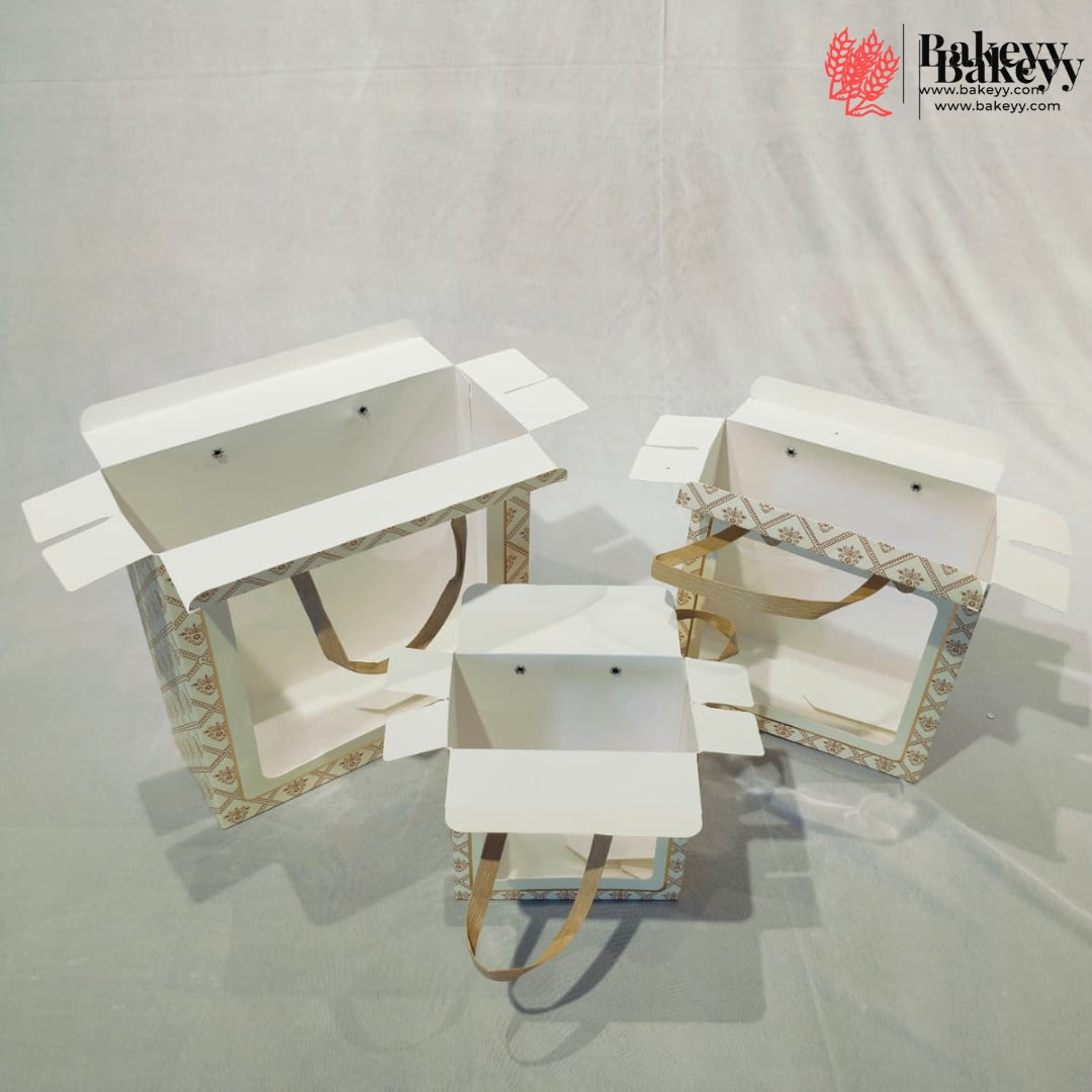 Three white bakery gift boxes with patterned sides and tan handles, displayed open on a light gray fabric backdrop. A bakery logo watermark is visible in the top right corner.