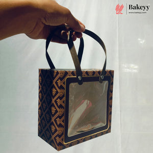 A hand holds a square bakery gift box with a clear front window and dark brown handles. The box has a dark brown and gold geometric pattern.