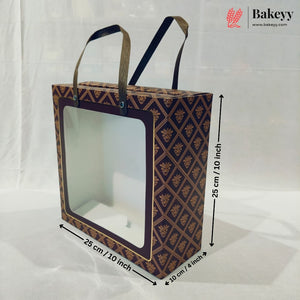 A square bakery gift box with a clear front window and two handles, shown with dimension arrows and labels indicating 25 cm by 25 cm by 10 cm. The box has a dark navy and gold geometric pattern.