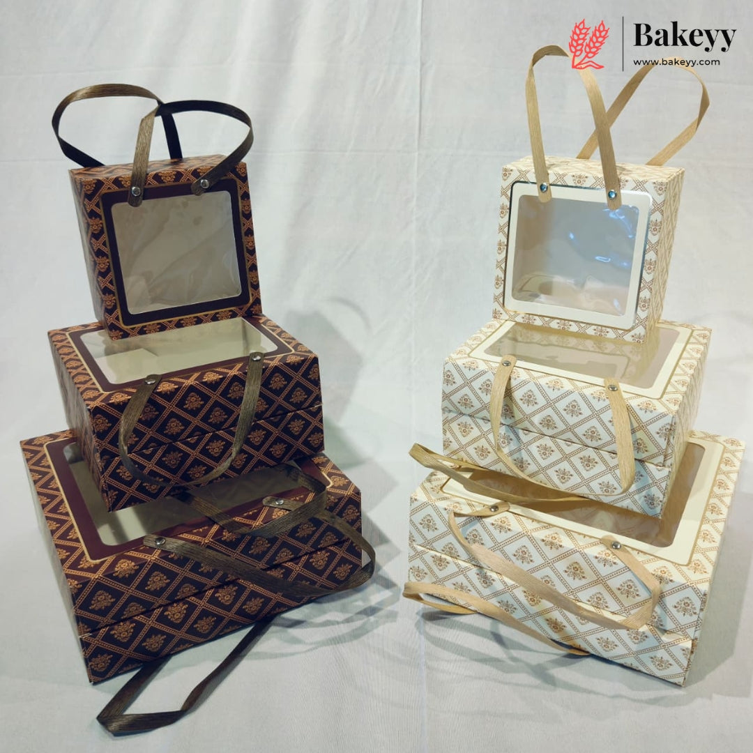 Two sets of decorative stacked gift boxes with clear window lids and ribbon handles, shown on a white fabric backdrop. One set is dark brown with a gold geometric pattern and dark brown ribbon, and the other is cream and gold with a matching geometric pattern and cream ribbon.