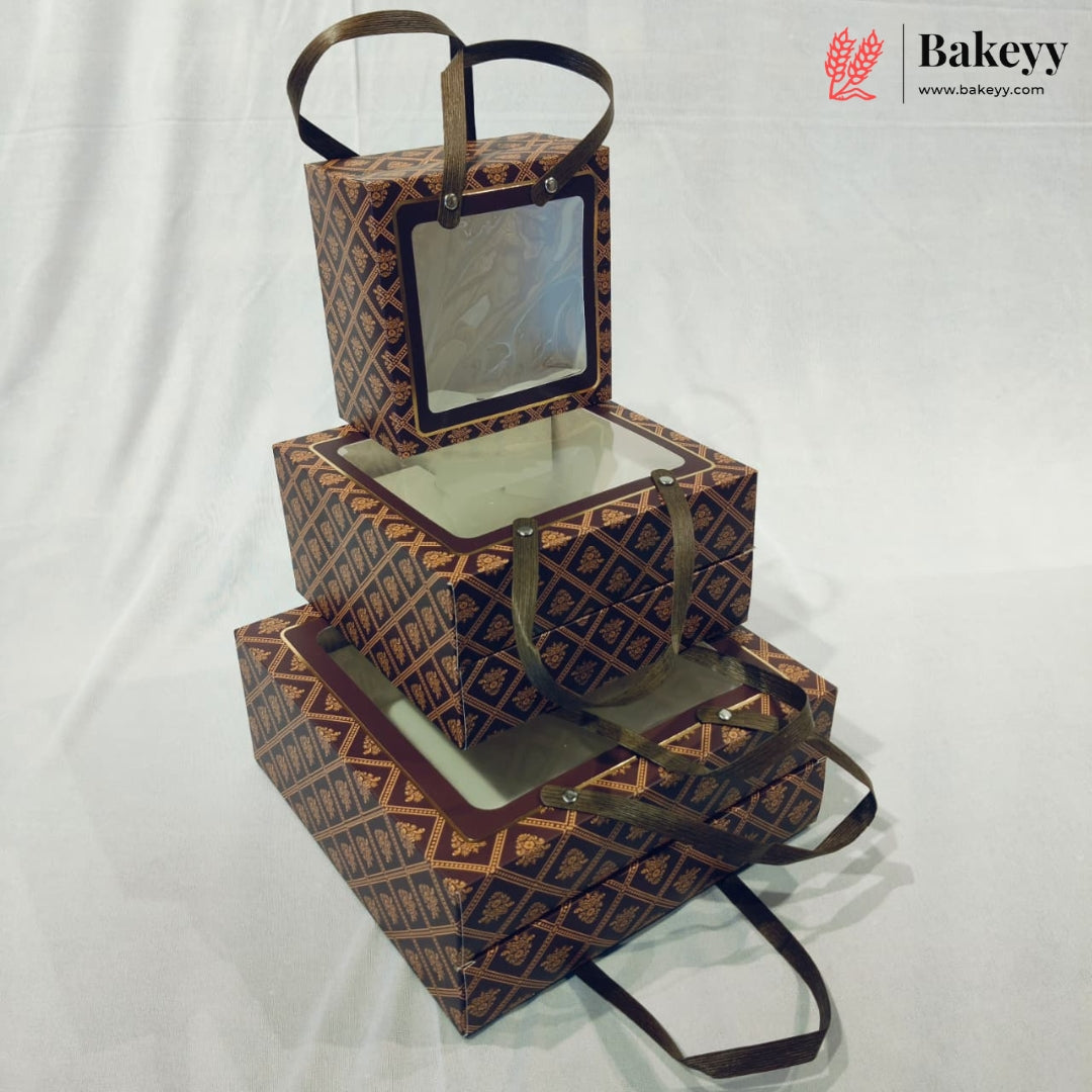 Three stacked rectangular bakery boxes with clear window lids and dark brown handles, featuring a repeating geometric pattern in brown and gold. The boxes are arranged on a white fabric backdrop, with a Bakeyy logo watermark in the top right corner.