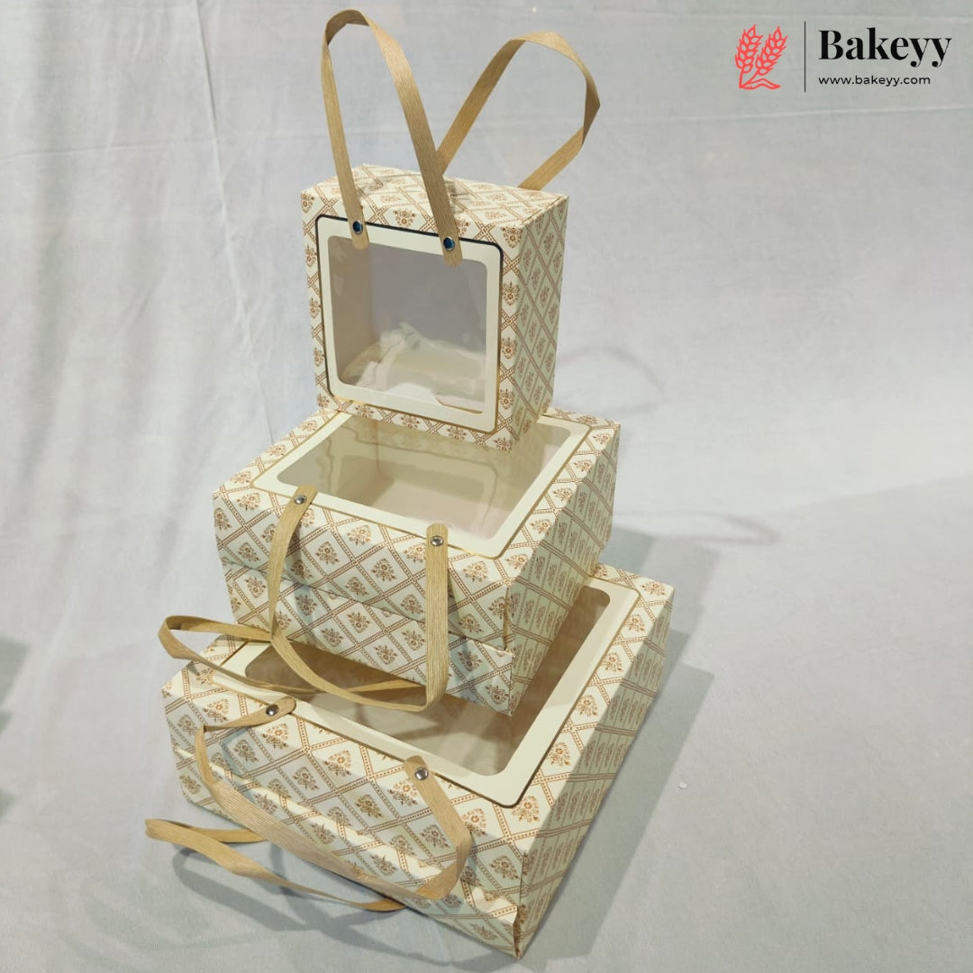 Three nested rectangular bakery boxes with clear window tops and tan ribbon handles, photographed on a light gray fabric backdrop. A Bakeyy logo watermark appears in the top right corner.