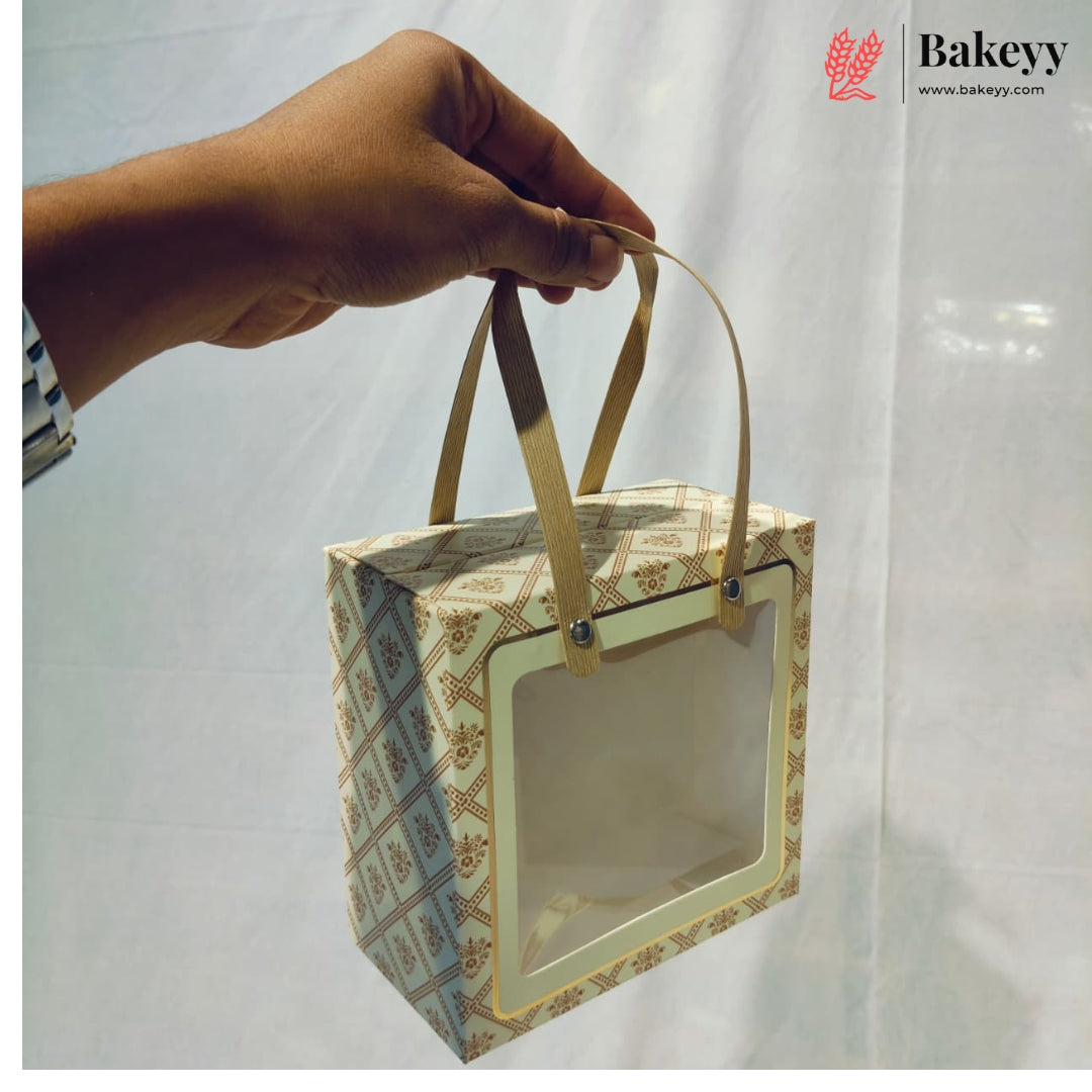 A hand holds a square bakery gift box with a clear front window and two tan handles. The box has a cream and gold geometric pattern.